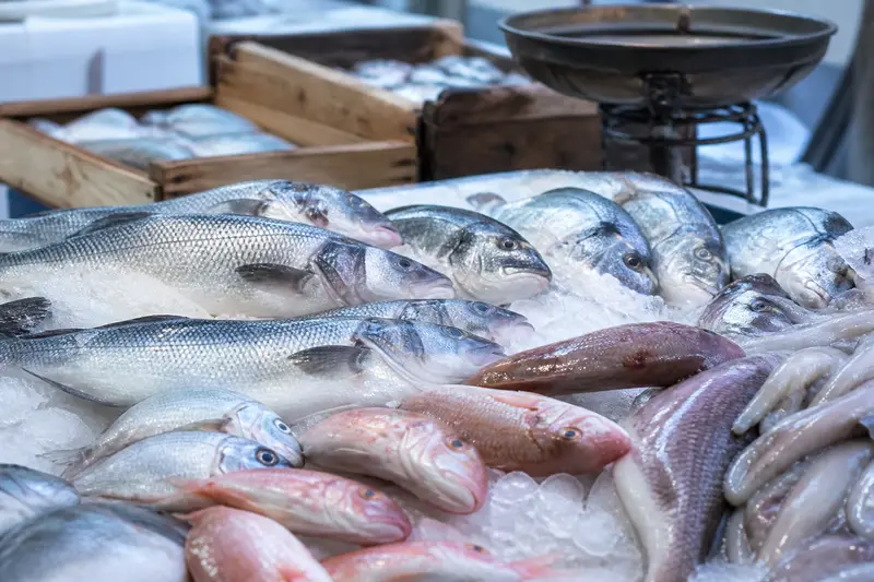 Étal de poissons frais sur glace au marché : loups, daurades et rougets, avec des cageots en bois derrière.